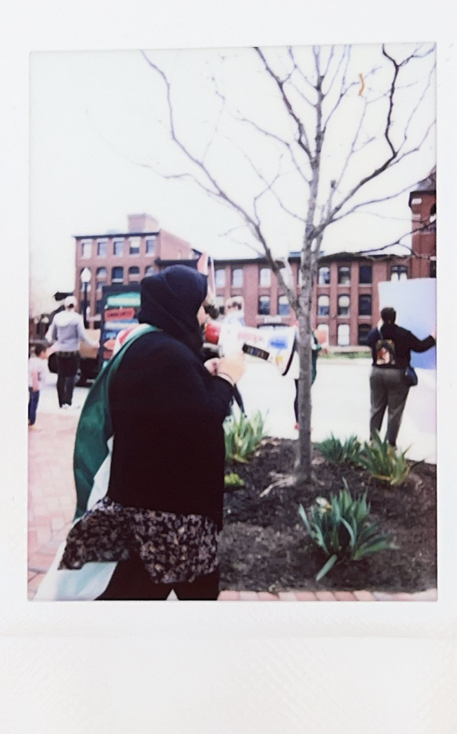 polaroid photo of protestors holding signs in front of a brick mill building. in the foreground, a woman in black with a Palestinian flag draped on her shoulders holds a megaphone.